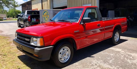 Bright red pickup truck parked in front of industrial buildings and other vehicles on a sunny day