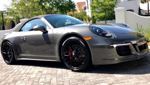 Silver Porsche 911 sports car parked on brick driveway with open driver door and trees in background