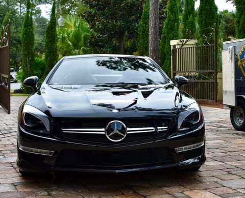 Black Mercedes-Benz sports car parked in driveway with brick pavement and cypress trees in background