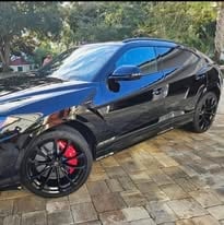 Black sports car with blue top and red wheels parked on brick driveway with trees in background