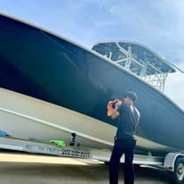 Person standing beside a large dark blue boat on a trailer under cloudy sky
